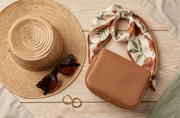 Mise en scène flat lay d'accessoires de mode tendance incluant chapeau en paille, lunettes écaille, foulard en soie et sac en cuir sur bois blanchi